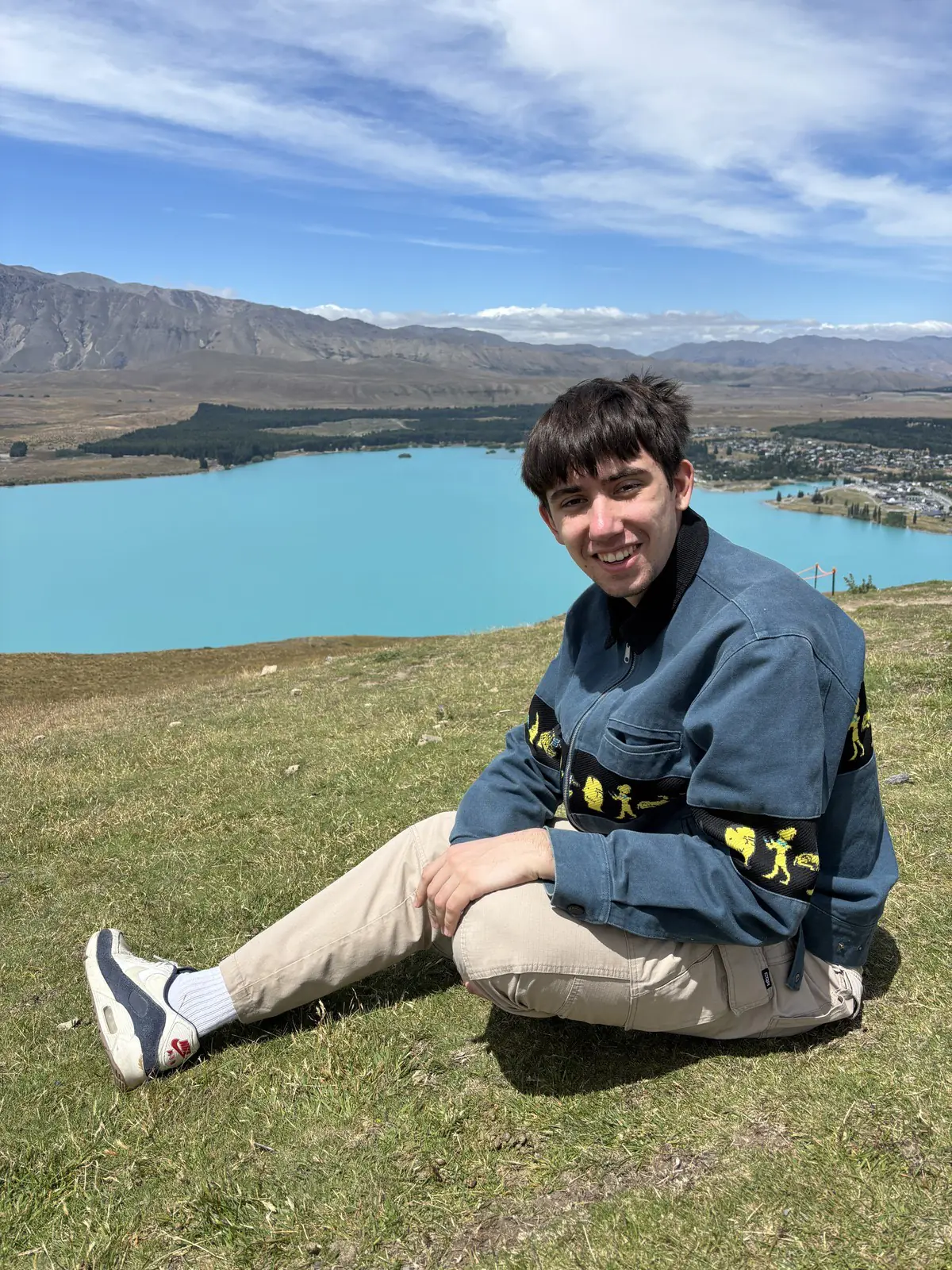 John sitting on the grass above a turquoise lake in New Zealand